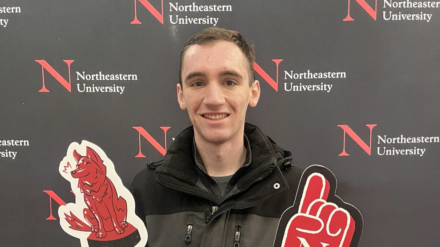 Ryan Mullin smiling in a black jacket in front of a Northeastern University step-and-repeat backdrop, between a cutout of the red husky mascot and a cutout of a red foam finger